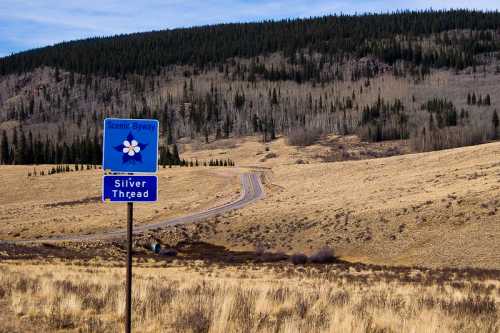 A scenic byway sign for Silver Thread stands beside a winding road through a grassy landscape and distant forested hills.