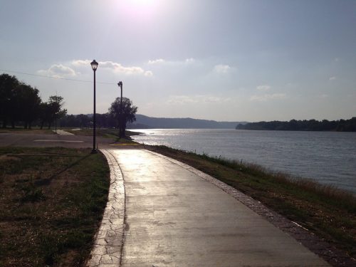 A serene riverside path with lampposts, sunlight reflecting on the water, and trees lining the shore.