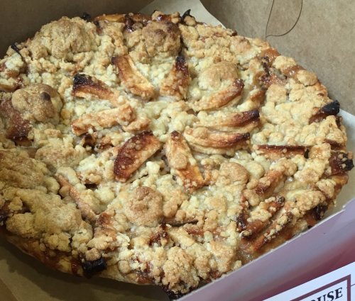 A freshly baked apple pie with a crumbly topping, displayed in a cardboard box.