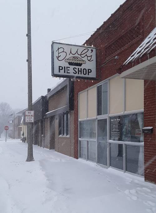 A snowy street scene featuring a sign for "Bob's Pie Shop" with snow covering the ground and buildings.
