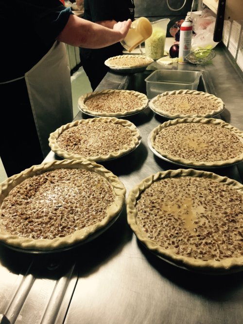 A kitchen scene with several unbaked pecan pies on a counter, as someone pours a filling into one of the pies.