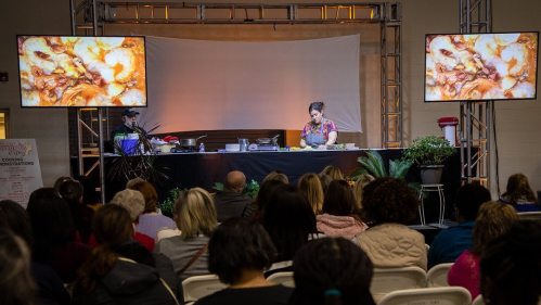 A cooking demonstration with a chef on stage, large screens displaying food, and an audience seated in front.