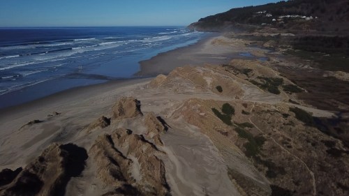 Aerial view of a sandy beach with gentle waves, rocky formations, and a coastal landscape under a clear blue sky.