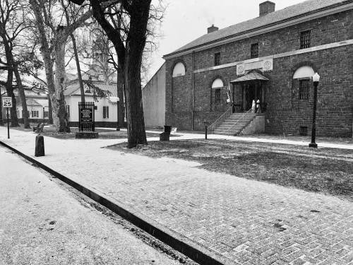 A snowy scene featuring historic buildings, trees, and a pathway, captured in black and white.