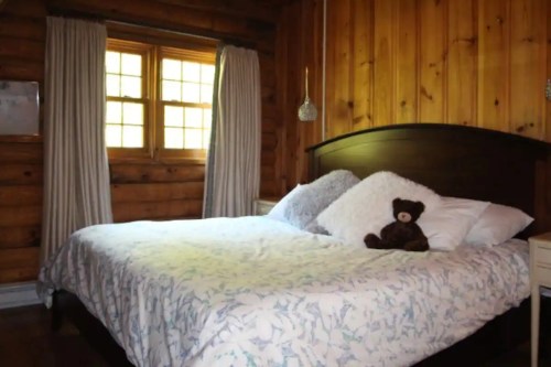 Cozy bedroom with a wooden interior, featuring a bed with pillows and a teddy bear on top. Natural light from a window.