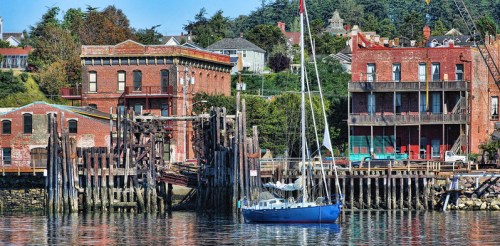 A blue sailboat docked near weathered brick buildings and wooden pilings by the water on a clear day.