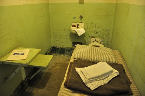 A small, sparse prison cell with a bed, sink, and a stack of folded blankets on the floor. Walls are painted green.