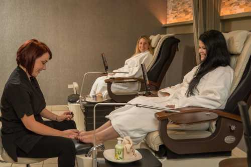 Two women in robes relax in spa chairs while a technician gives a pedicure to one of them.