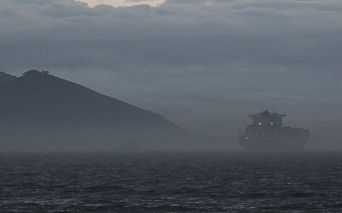 A large ship is silhouetted against a misty landscape, with dark clouds and a distant hill in the background.