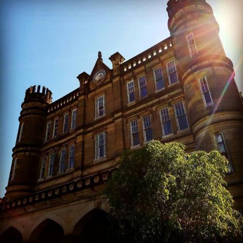 Historic building with towers and a clock, bathed in sunlight, surrounded by greenery against a clear blue sky.