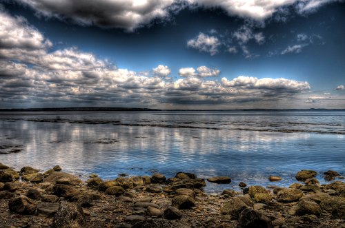 A serene lakeside view with rocky shore, calm water reflecting clouds, and a dramatic sky above.