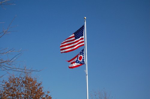 Two flags flying on a pole: the American flag above and the Ohio state flag below, against a clear blue sky.