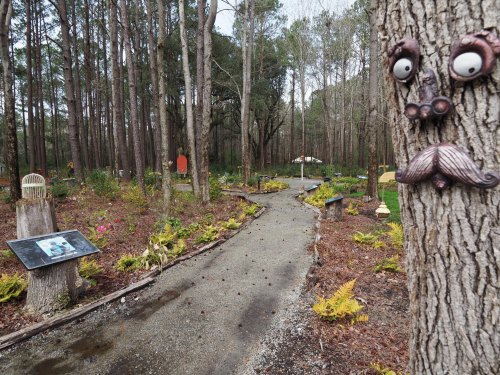 A whimsical garden path winds through trees, featuring quirky sculptures and art installations among lush greenery.