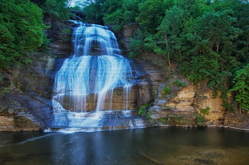 A serene waterfall cascading down rocky cliffs, surrounded by lush green trees and reflecting in a calm pool below.