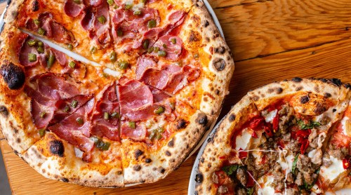 Two pizzas on a wooden table: one topped with ham and green peppers, the other with sausage and cheese.