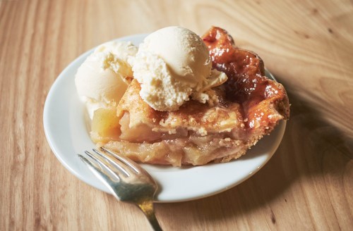 A slice of apple pie topped with a scoop of vanilla ice cream on a white plate, with a fork beside it.