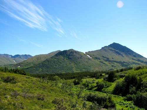 A scenic view of green hills and mountains under a clear blue sky, with a few patches of snow visible on the peaks.