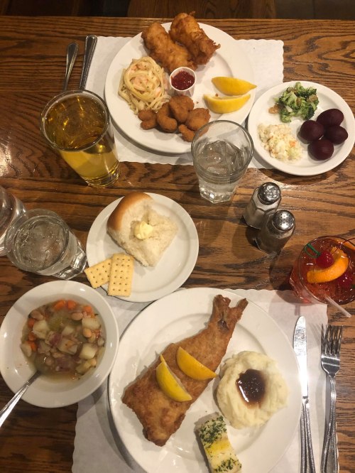 A table set with two plates of fried fish, sides of pasta, salad, mashed potatoes, and drinks, including water and a cocktail.