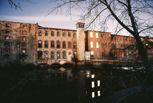 Abandoned industrial building by a river at dusk, with reflections in the water and trees in the foreground.