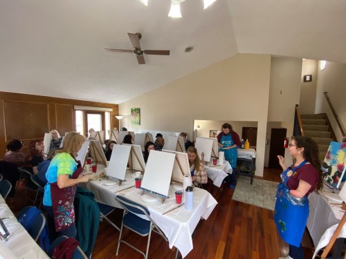 A group of people in an art class, seated at easels with blank canvases, listening to an instructor in a spacious room.