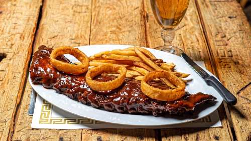 A plate of BBQ ribs topped with onion rings, served with French fries and a glass of beer on a rustic wooden table.