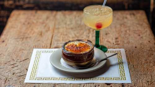 A bowl of creamy dessert with a caramelized top next to a colorful cocktail on a wooden table.
