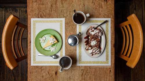 A wooden table with two coffee cups, a slice of cheesecake with whipped cream, and a chocolate dessert with toppings.