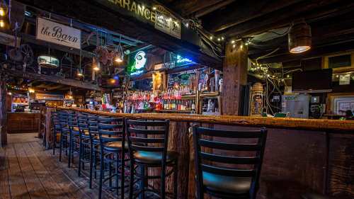 A rustic bar interior with wooden beams, a long counter, and colorful lights illuminating shelves of liquor bottles.