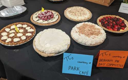A display of various pies on a table, labeled with signs from Park Café and the Braham Pie Day Committee.