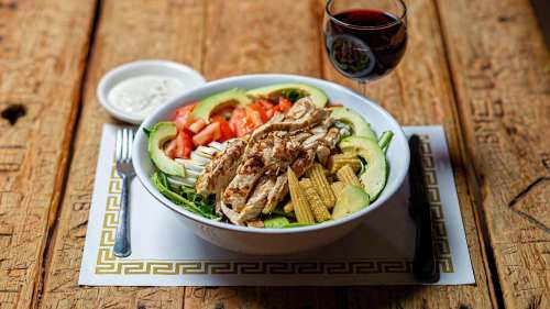 A colorful salad with grilled chicken, avocado, tomatoes, and corn, served with a side of dressing and a glass of red wine.
