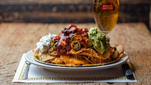 A plate of nachos topped with cheese, jalapeños, guacamole, sour cream, and salsa, alongside a glass of beer.
