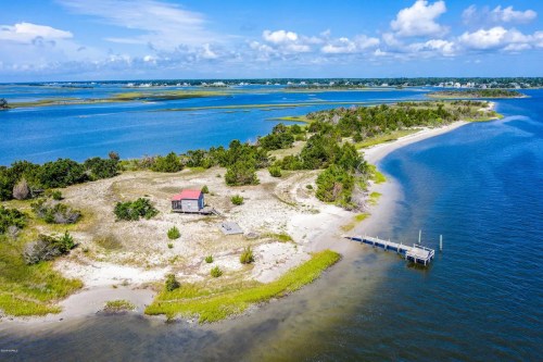 Aerial view of a coastal landscape featuring a small house, sandy shore, and a dock by the water under a blue sky.