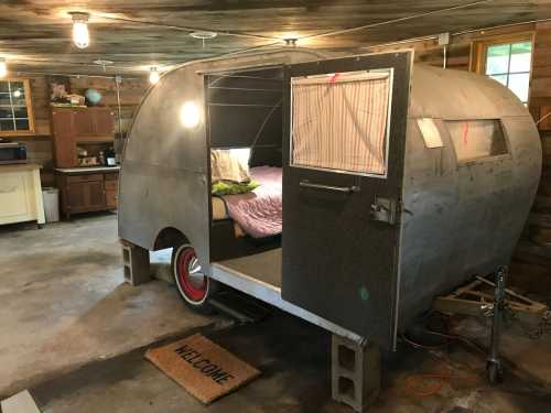 A vintage camper trailer inside a garage, featuring a bed and a welcome mat at the entrance.