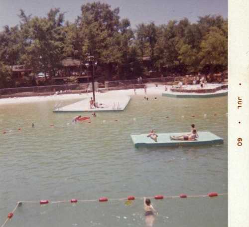 A vintage photo of a swimming pool area with people swimming, lounging on rafts, and enjoying the sun.