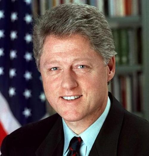 Portrait of a man with gray hair, smiling, wearing a suit, with an American flag in the background.