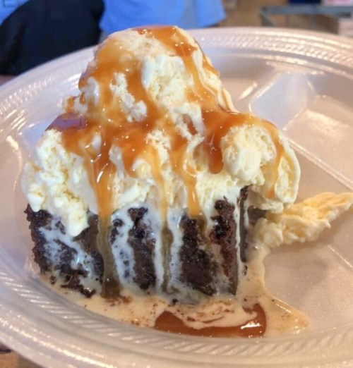 A slice of chocolate cake topped with melting vanilla ice cream and drizzled with caramel sauce on a paper plate.