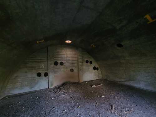 Interior of a dimly lit concrete bunker with numbered walls and dark circular marks, featuring a dirt floor.