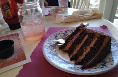 A slice of chocolate cake on a plate next to a jar of pink drink and a container of ketchup on a table.