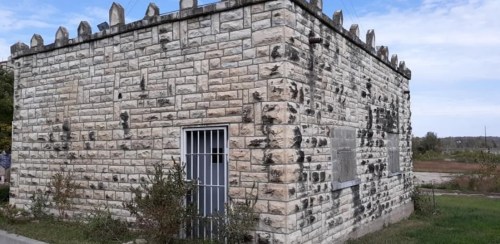 A small, weathered stone building with a barred window and a flat roof, surrounded by overgrown grass and shrubs.