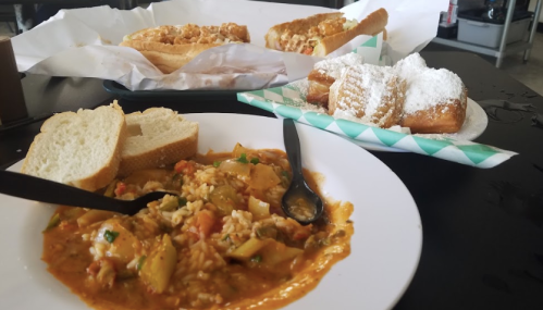 A plate of gumbo with rice, slices of bread, and a dessert of powdered sugar beignets on a table.