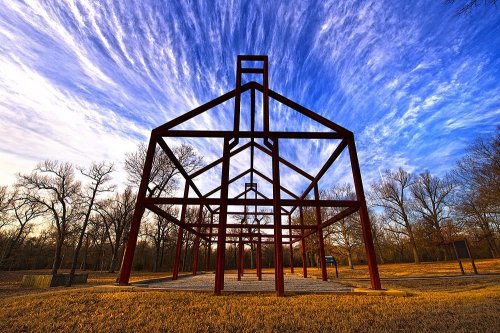 A skeletal metal structure stands in a grassy area under a vibrant sky with wispy clouds and bare trees.