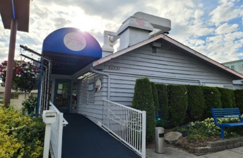 A charming restaurant with a blue awning, surrounded by greenery and flowers, under a partly cloudy sky.