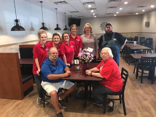 A group of eight people, including staff and customers, pose together at a restaurant table with flowers.