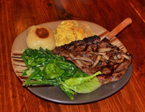 A plate of grilled steak with onions, greens, a roll, and macaroni and cheese on a wooden table.