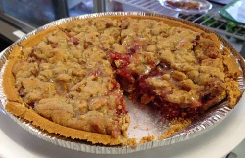 A pie with a crumbly topping and a slice removed, revealing a cherry filling inside a golden crust.
