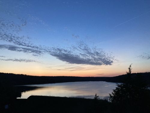 A serene lake at sunset, reflecting colorful clouds and a twilight sky, surrounded by trees and hills.
