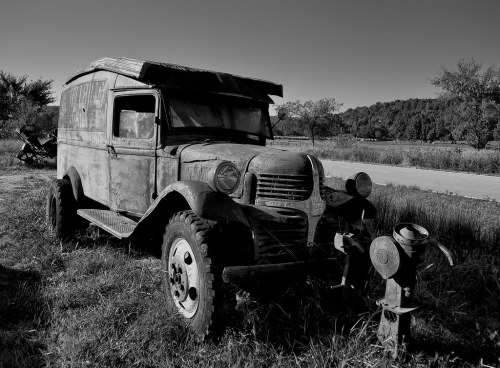 An old, rusty truck parked in a grassy area, with trees and a road in the background, captured in black and white.