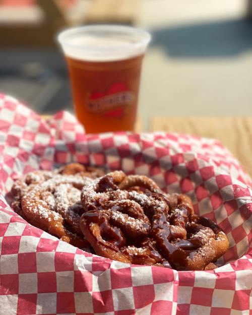 A basket of warm, powdered sugar-covered funnel cake with chocolate drizzle, next to a cup of beer.