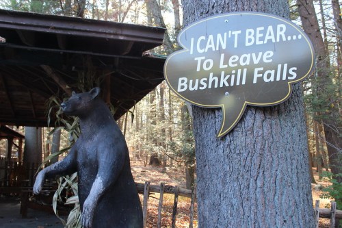 A bear statue next to a sign that says, "I CAN'T BEAR... To Leave Bushkill Falls," in a wooded area.