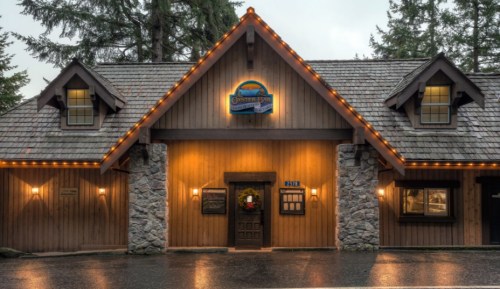 A cozy wooden restaurant with stone accents, warmly lit, surrounded by trees on a rainy day.
